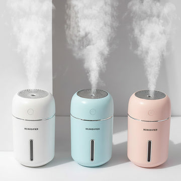 Three humidifiers in white, blue, and pink emitting steam on a light gray background.